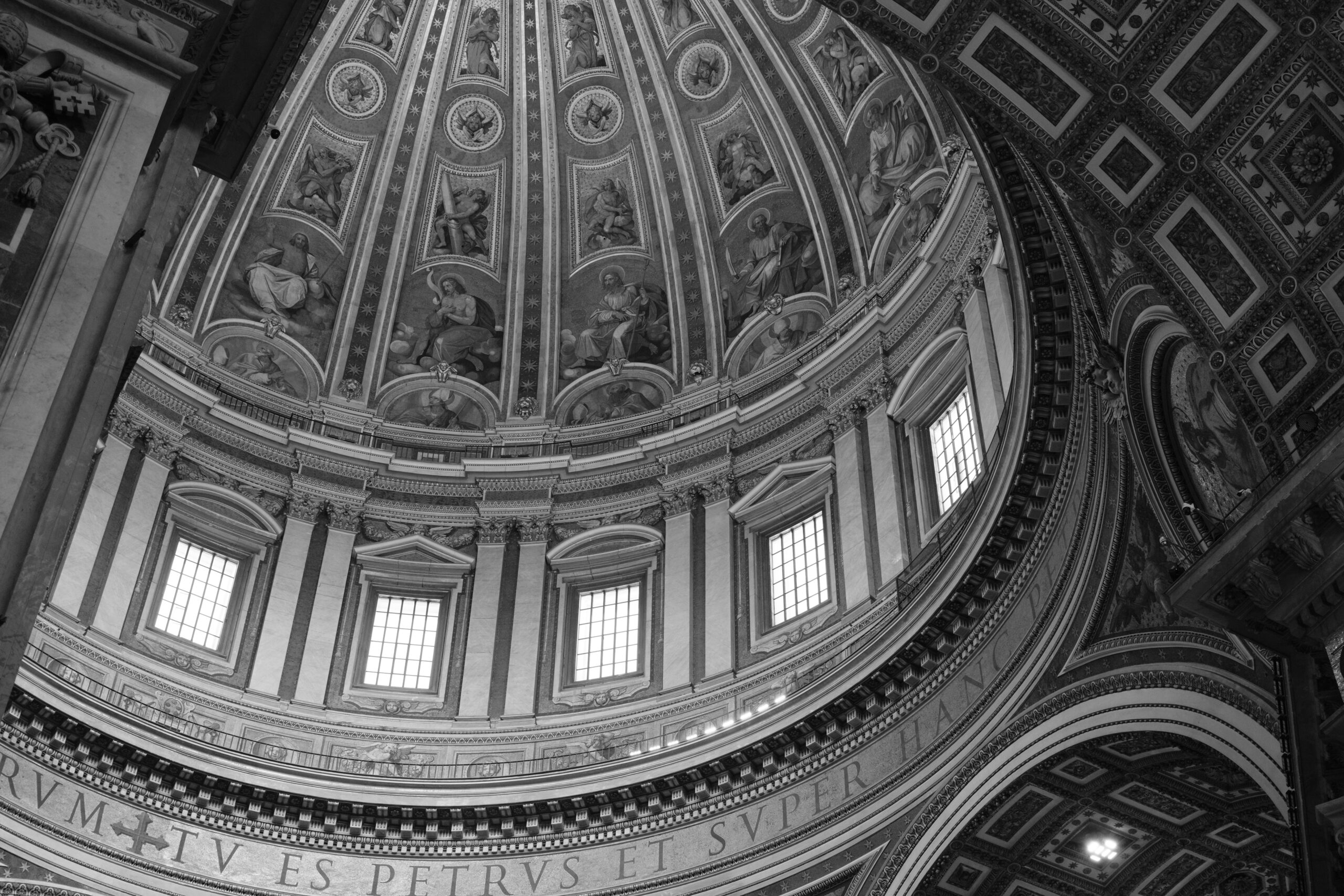 Black and white photograph of the interior dome and surrounding arches of St. Peter's Basilica in Vatican City. The image captures the intricate frescoes and detailed architectural designs, including classical columns and decorative moldings. Latin inscriptions run along the circular band beneath the dome, bathed in natural light filtering through the dome's windows, enhancing the majestic and serene atmosphere of this iconic religious site.